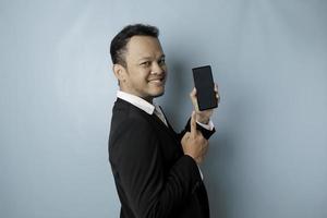 A portrait of a happy Asian businessman is smiling and holding his smartphone showing copy space on it's screen wearing black suit isolated by a blue background photo