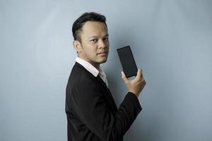 un joven hombre de negocios asiático con traje negro se siente serio y se enfoca sosteniendo un teléfono inteligente y mostrando el espacio de copia en su pantalla en la mano. foto
