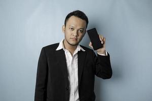 un joven hombre de negocios asiático con traje negro se siente serio y se enfoca sosteniendo un teléfono inteligente y mostrando el espacio de copia en su pantalla en la mano. foto