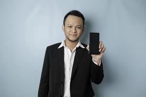 A portrait of a happy Asian businessman is smiling and holding his smartphone showing copy space on it's screen wearing black suit isolated by a blue background photo