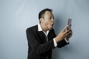Surprised Asian businessman wearing black suit holding his smartphone, isolated by blue background photo