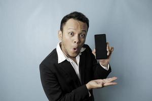 sorprendido hombre de negocios asiático con un traje negro sosteniendo su teléfono inteligente y mostrando espacio de copia en su pantalla, aislado por un fondo azul foto