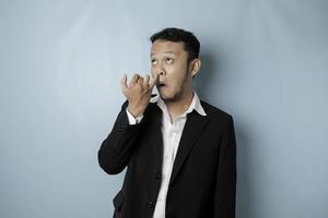 un hombre asiático vestido con traje negro se está recogiendo la nariz aislado de fondo azul. foto