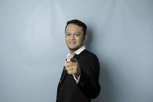 Portrait of young Asian businessman in black suit pointing index finger at camera, posing isolated over blue background. Cheerful smiling guy picking, choosing and indicating photo