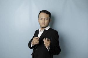 Portrait of young Asian businessman in black suit pointing index finger at camera, posing isolated over blue background. Cheerful smiling guy picking, choosing and indicating photo