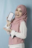 A portrait of a young Asian Muslim woman wearing pink hijab is smiling while showing her smartphone screen photo