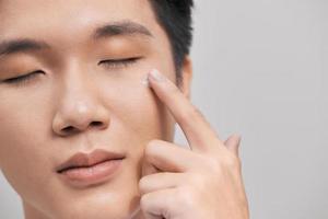 Young handsome man with moisturizer on his face, isolated on gray background. Skin care concept photo