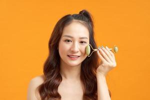 hermosa chica asiática desnuda masajeando la cara con un rodillo de jade aislado en naranja amarillo foto