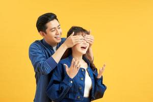 Photo of happy man smiling and covering woman's eyes standing behind her isolated over yellow background