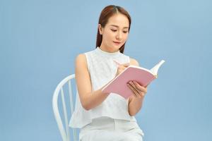 A young asian woman sitting and writing her notes isolated over blue background photo