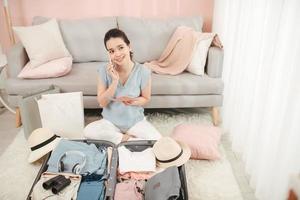 young woman talking with friend on mobile phone holding passport packing travel suitcase organizing clothes and things in summer holiday trip and wanderlust concept