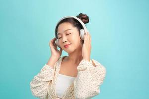 Portrait of a cheerful stylish young woman standing isolated over blue background, listening to music with headphones, dancing photo