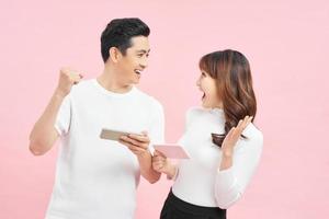 Beautiful couple wearing white t-shirts standing isolated over pink background, playing games on mobile phone. photo