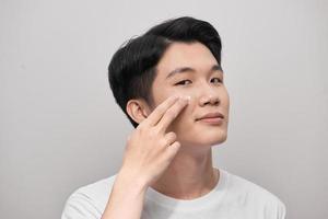 Skin care. Portrait of handsome young man applying cream to his face, isolated on white background photo