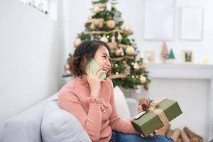 Technology. Communication. Christmas. Young woman is holding a gift box, talking on the mobile phone and smiling while sitting near the Xmas tree at home photo