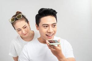 Portrait of a young couple using mobile phones while standing together over gray background, curious woman looking at mans phone photo