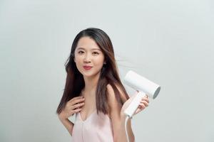 Attractive cheerful asian girl blows dry her hair with hairdryer, over white photo