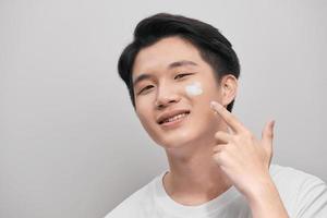 Skin care. Portrait of handsome young man applying cream to his face, isolated on white background photo