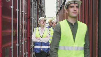 Group of multiracial workers in safety uniforms and hardhats walk in a deep row and inspect shipping cargo at nook of container stack, goods logistic transport industry, and professional duty labor. video