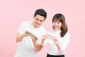 Two cheerful friends standing isolated over pink background, playing games on mobile phone. photo