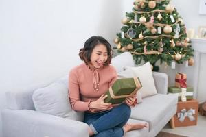 A young woman holds a gift near the Christmas tree. The girl smiles sitting with a gift for Christmas photo