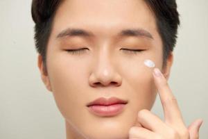 Portrait of satisfied young man applying facial cream isolated over white background photo