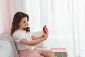 Attractive smiling young asian woman wearing casual clothes sitting on a couch, taking a selfie photo