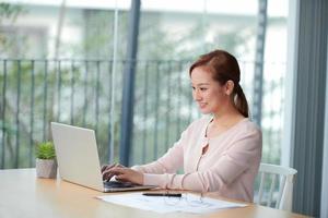 Beautiful young woman in casual clothing using laptop and smiling while working indoors photo