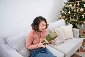 Beautiful asian woman in casual sweater holding gift near decorated christmas tree, greeting card concept photo