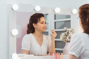 Happy attractive young female applying cosmetics on face near cosmetic table in morning photo