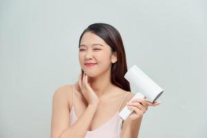 Attractive cheerful asian girl blows dry her hair with hairdryer, over white photo