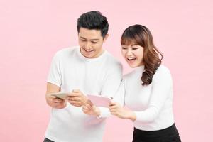 Image of excited couple man and woman smiling while both using mobile phones isolated over pink background photo