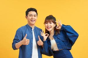 Excited beautiful asian couple shows thumb up gesture agree to do something and collaborate against orange wall, did great job, show approval, like idea. Everything will be OK. photo