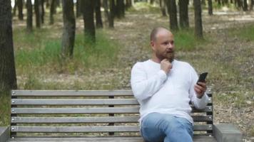 un hombre con una camisa blanca con barba en el parque en un banco hablando por teléfono video