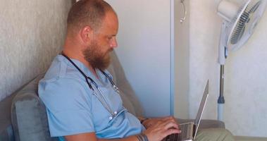 A man with a beard in a doctor's suit is typing on a laptop in a rest room video
