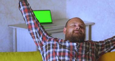 Happy man in a plaid shirt with a beard sits on the couch in the room video