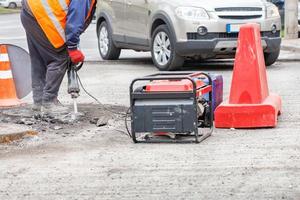 A worker using an electric jackhammer and a gasoline generator repairs old asphalt on a fenced section of the road. photo