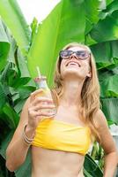 joven mujer caucásica en traje de baño amarillo bebiendo bebidas de pie en hojas de palmeras tropicales foto