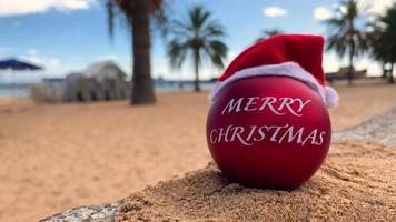 weihnachten, rote bombe in santa's hat am exotischen strand mit palmen und blauem himmel im hintergrund mit wolken. Hawaii, Kanarische Inseln, Bali, Thailand. Frohe Weihnachten aus dem Paradies. video