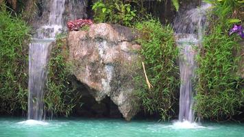 una cascada artificial que desemboca en la superficie del agua azul. La estructura de la cascada consiste en árboles naturales y flores que se ven brillantes durante el día. video