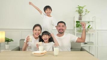 una familia tailandesa asiática saludable, niños pequeños y padres jóvenes beben leche blanca fresca en un vaso y pan de alegría juntos en una mesa de comedor en la mañana, bienestar nutrición hogar desayuno comida estilo de vida. video
