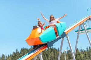 Girls are going down the hill in a mountain coaster with outstretched arms. Mountain trees in background photo