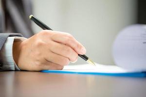 Workplace closeup person professional businesswoman sitting at desk hold pen signing or signature contract paper. Employee woman writing agreement document on paperwork form corporate at work office photo