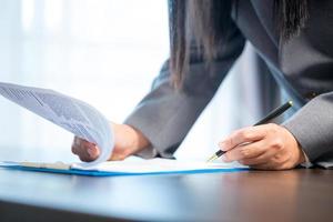 Workplace closeup person professional businesswoman sitting at desk hold pen signing or signature contract paper. Employee woman writing agreement document on paperwork form corporate at work office photo