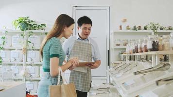 Asian male shopkeeper describes natural organic products to woman customer in refill store, zero-waste and plastic-free grocery, eco environment-friendly, sustainable lifestyles with a reusable shop. video