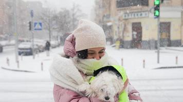 una mujer abraza a un pequeño perro blanco afuera en la nieve, ambos con abrigos hinchados video