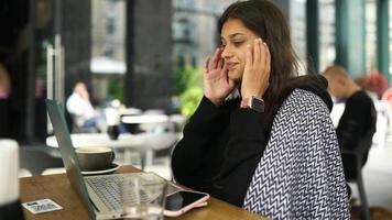 Woman at a coffee shop table uses laptop and rubs temples in distress video