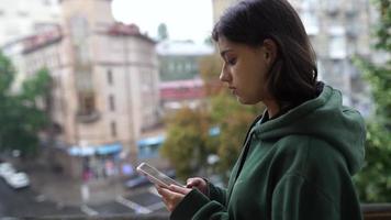 giovane donna nel felpa con cappuccio usi inteligente Telefono al di fuori come esso piove video