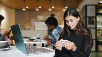 mulher no café olha para seu telefone inteligente enquanto está sentado em uma mesa com seu laptop video