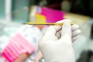 Doctor eyes view looking numbers on small medical thermometer in his hands with blurry a sick newborn baby sleeping in a baby bed. photo